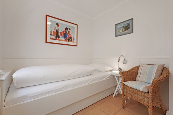 Un lit simple avec une literie blanche, une chaise en osier avec un coussin et deux tableaux encadrés accrochés au mur.