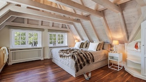 Un dormitorio con techo inclinado, vigas de madera, cama con una manta de piel, mesita de noche y radiador.