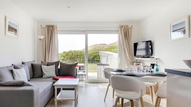 Un salon moderne avec un canapé gris, une table à manger avec des chaises, un téléviseur à écran plat et une grande fenêtre avec des rideaux.