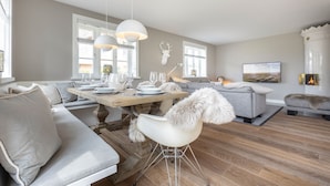 Ein modernes Wohnzimmer mit Essbereich, grauen Sofas, einem weißen Sessel mit Felldecke, Holzböden und einem aufgehängten Hirschkopf an der Wand.