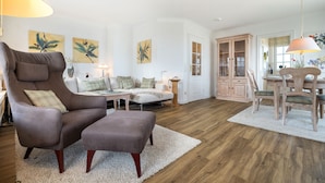 Un salon spacieux avec un grand fauteuil, une petite table et un coin repas comprenant une table et des chaises en bois. Des tableaux ornent les murs et une commode se trouve au fond de la pièce.