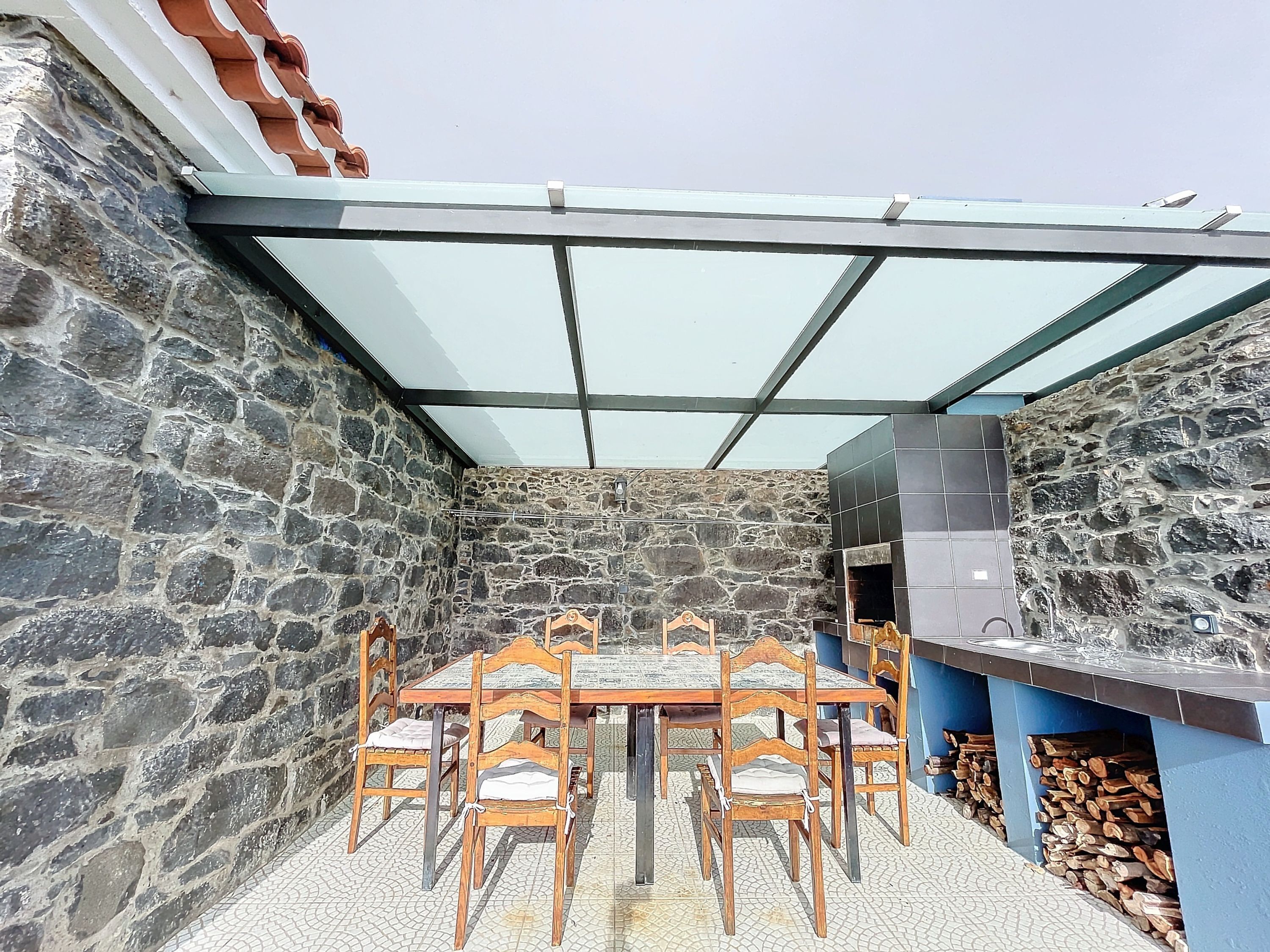 A stone-walled patio with a glass roof, a tiled floor, and a wooden table with chairs.