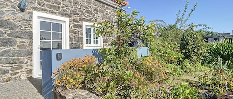 A stone building with a red-tiled roof, a blue door, and a garden with various plants and flowers.