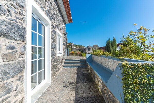 A stone building with a white window, a cobblestone path, and a blue wall with greenery. - Casa da Horta by Atlantic Holiday (Santa Cruz)