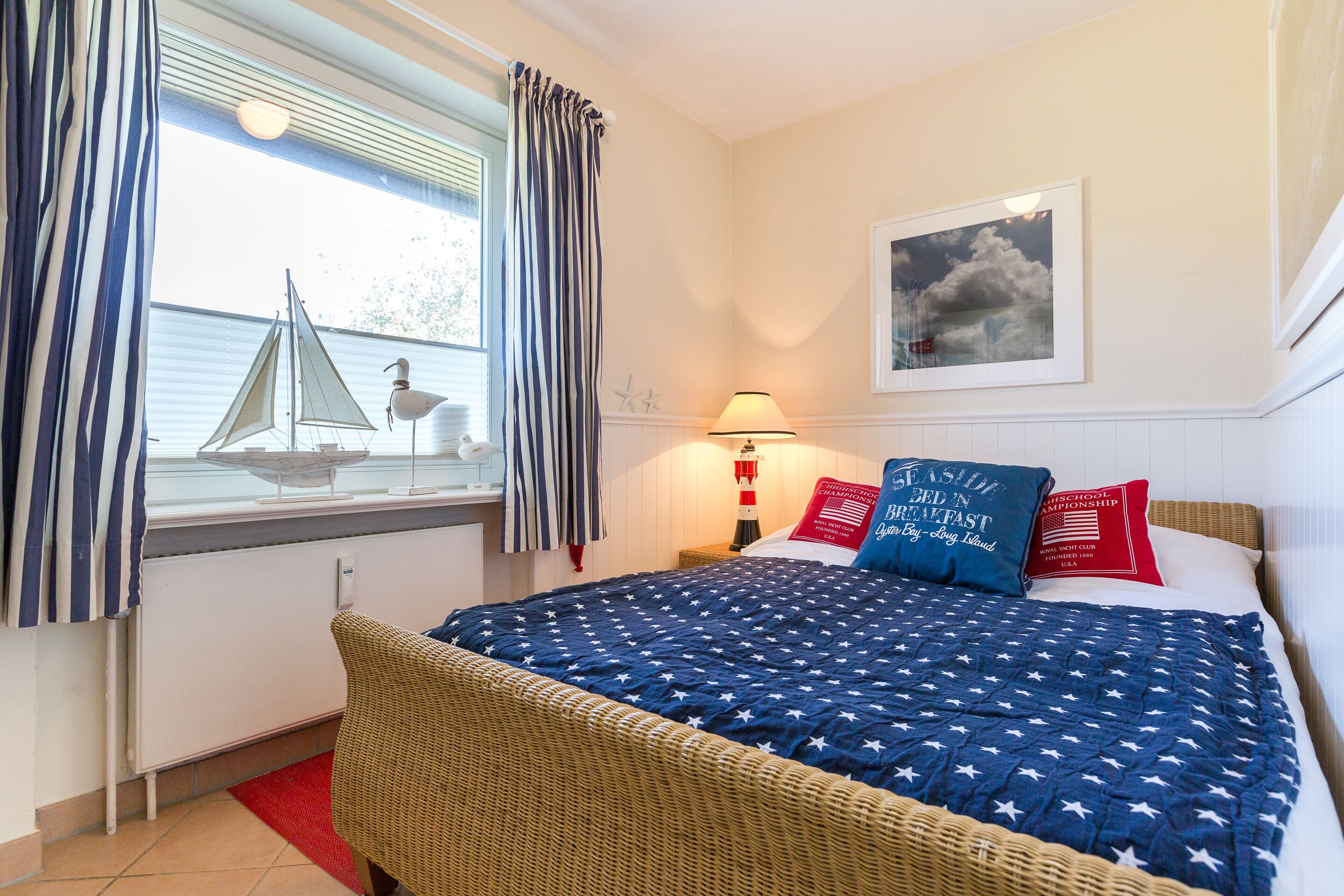 Een slaapkamer met een bed, een raam met gordijnen, een lamp, een lijst met een afbeelding en een klein model zeilboot.