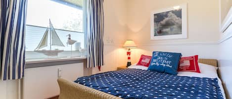 Een slaapkamer met een bed, een raam met gordijnen, een lamp, een lijst met een afbeelding en een klein model zeilboot.