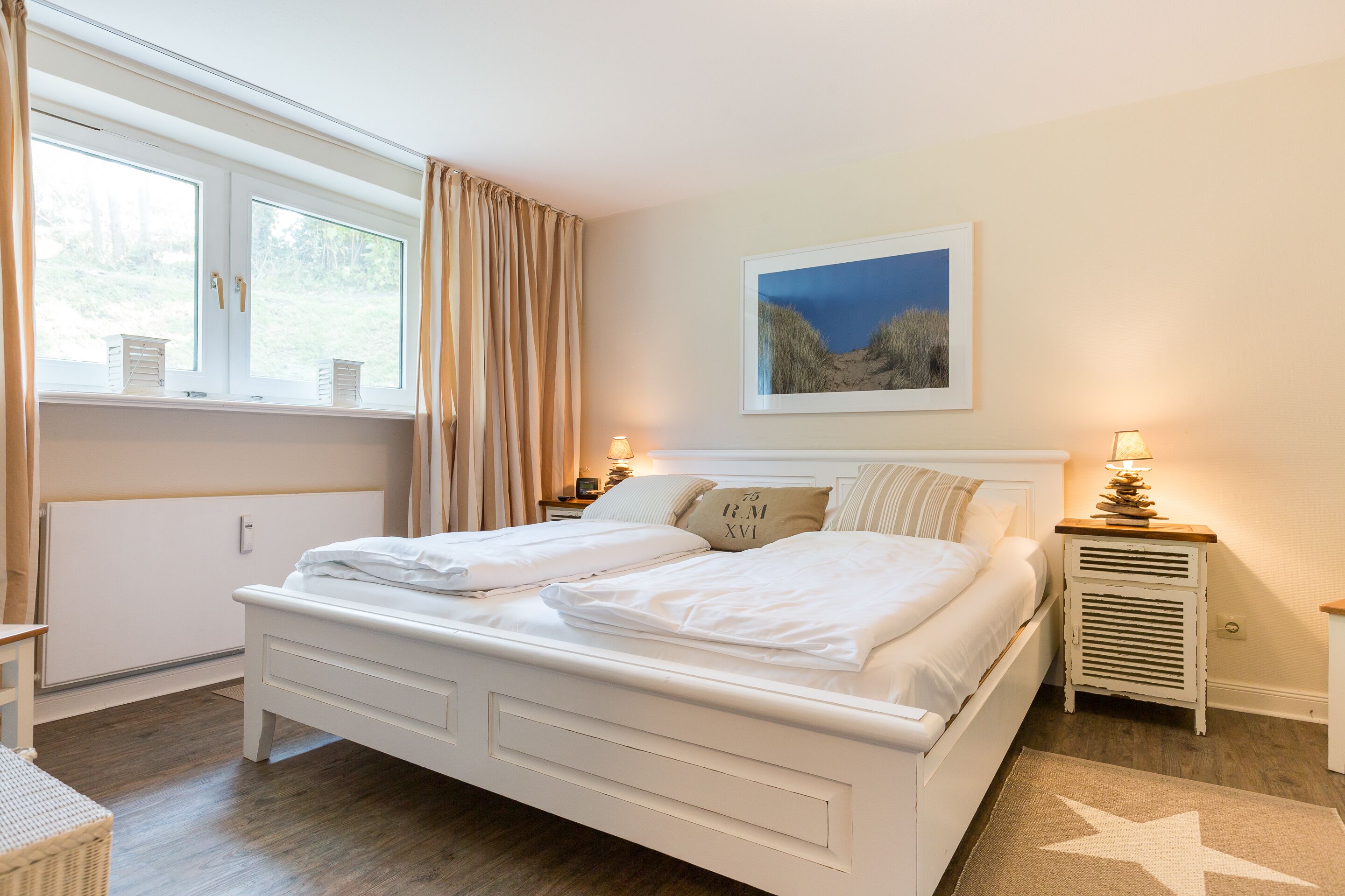 Ein Schlafzimmer mit einem großen Bett, einem Fenster mit Vorhängen, einem Nachttisch mit Lampe und einem gerahmten Bild an der Wand.
