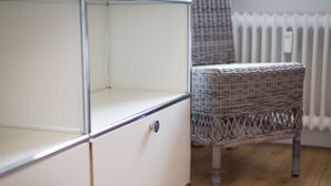 Un meuble blanc avec une poignée en métal, une chaise en osier et un radiateur à l’arrière-plan.