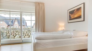 A bedroom with a bed, a window with curtains, a framed picture on the wall, and a view of buildings outside. - Completely ground floor 3-room apartment near the beach. (Wenningstedt-Braderup (Sylt))