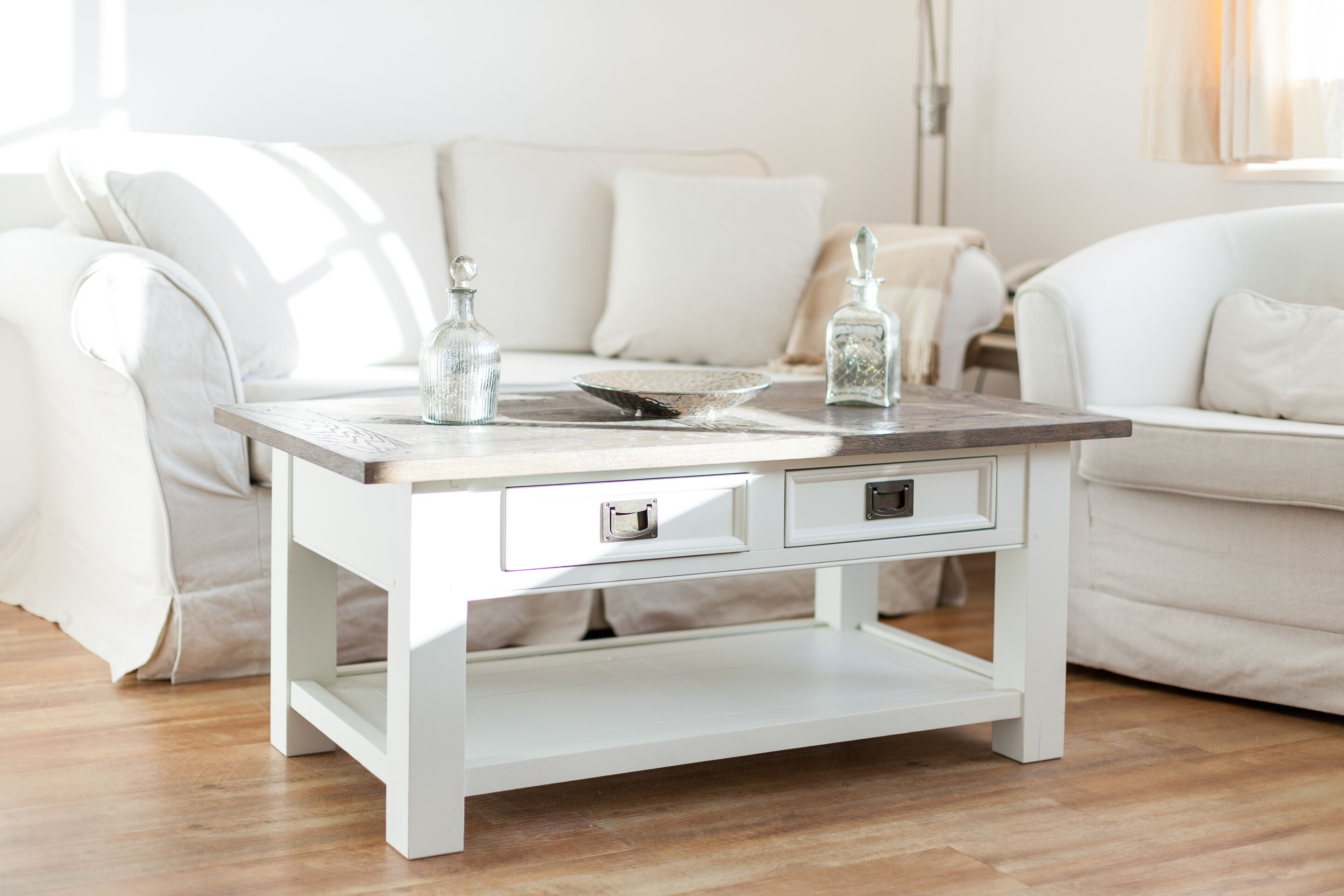 A white wooden coffee table with drawers, placed in a living room with a white sofa and a lamp.