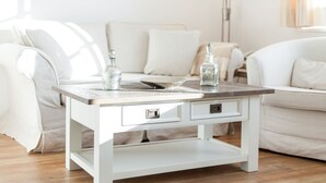 Una mesa de centro de madera blanca con cajones, ubicada en una sala de estar con un sofá blanco y una lámpara.
