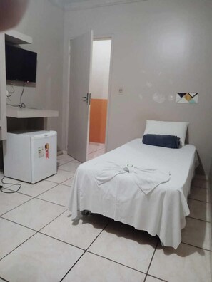Une chambre avec un lit, un petit réfrigérateur, un téléviseur fixé au mur et une porte donnant sur une autre pièce.