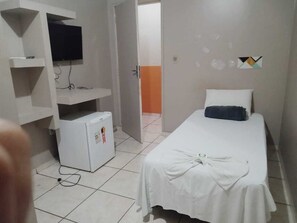 Une petite chambre propre, avec un lit, un téléviseur posé sur une étagère et un petit réfrigérateur.