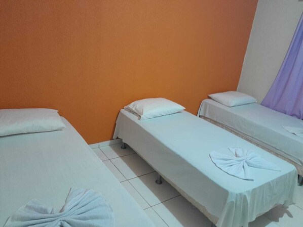 Quarto com três camas de solteiro, cada uma com colchão e travesseiro brancos, e uma toalha bem dobrada na cama em primeiro plano.
