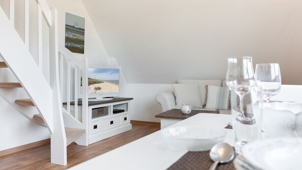 A modern, white-themed room with a staircase, a dining area with a table set for two, and a sofa with cushions. - 2-raum-appartement im OG und DG, ca. 43qm² Nutzfläche für bis zu 2 Personen (Wenningstedt-Braderup (Sylt))
