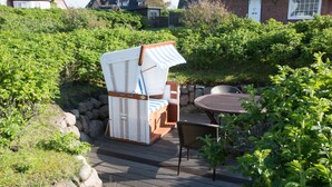 A wooden deck with a beach chair and a table, surrounded by greenery and a stone wall. - 2-raum-appartement im OG und DG, ca. 43qm² Nutzfläche für bis zu 2 Personen (Wenningstedt-Braderup (Sylt))