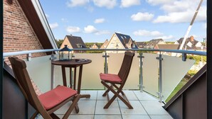 A balcony with two wooden chairs and a small round table, overlooking a residential area with houses and a clear sky. - Fantastisch Strandnahe Lage & Beach-style Wohlfühl-atmosphäre (Sylt / OT Westerland)