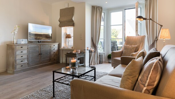 Een woonkamer met een bank, fauteuil en salontafel. Er is een televisie, een open haard en een raam met gordijnen.