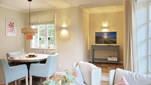 A dining area with a table and chairs, a sofa with decorative pillows, a wall-mounted TV, and a window with curtains.