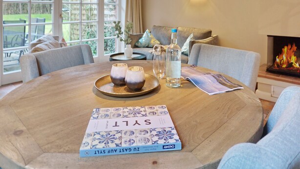 A round wooden table with a book titled "ZUR GAST AUF SYLL" and two glasses on a tray, a bottle of water, and a menu on the table.