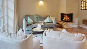 A cozy living room with a grey sofa, a white coffee table, and two grey armchairs. A fireplace with a visible fire adds warmth to the room.