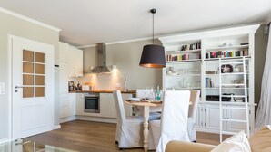 A modern kitchen with white cabinetry, a dining table with chairs, and a bookshelf filled with books and decorative items. - The most beautiful views! (Wenningstedt-Braderup (Sylt))