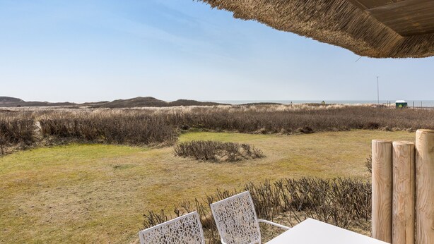 Un área de estar al aire libre con techo de paja, con vista a un paisaje cubierto de hierba y vegetación escasa, con colinas al fondo.