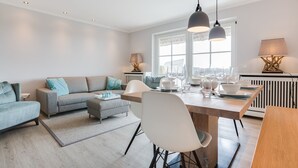 Un salón moderno con un sofá gris, una mesa de comedor con cristalería y vistas a la ciudad a través de grandes ventanales.