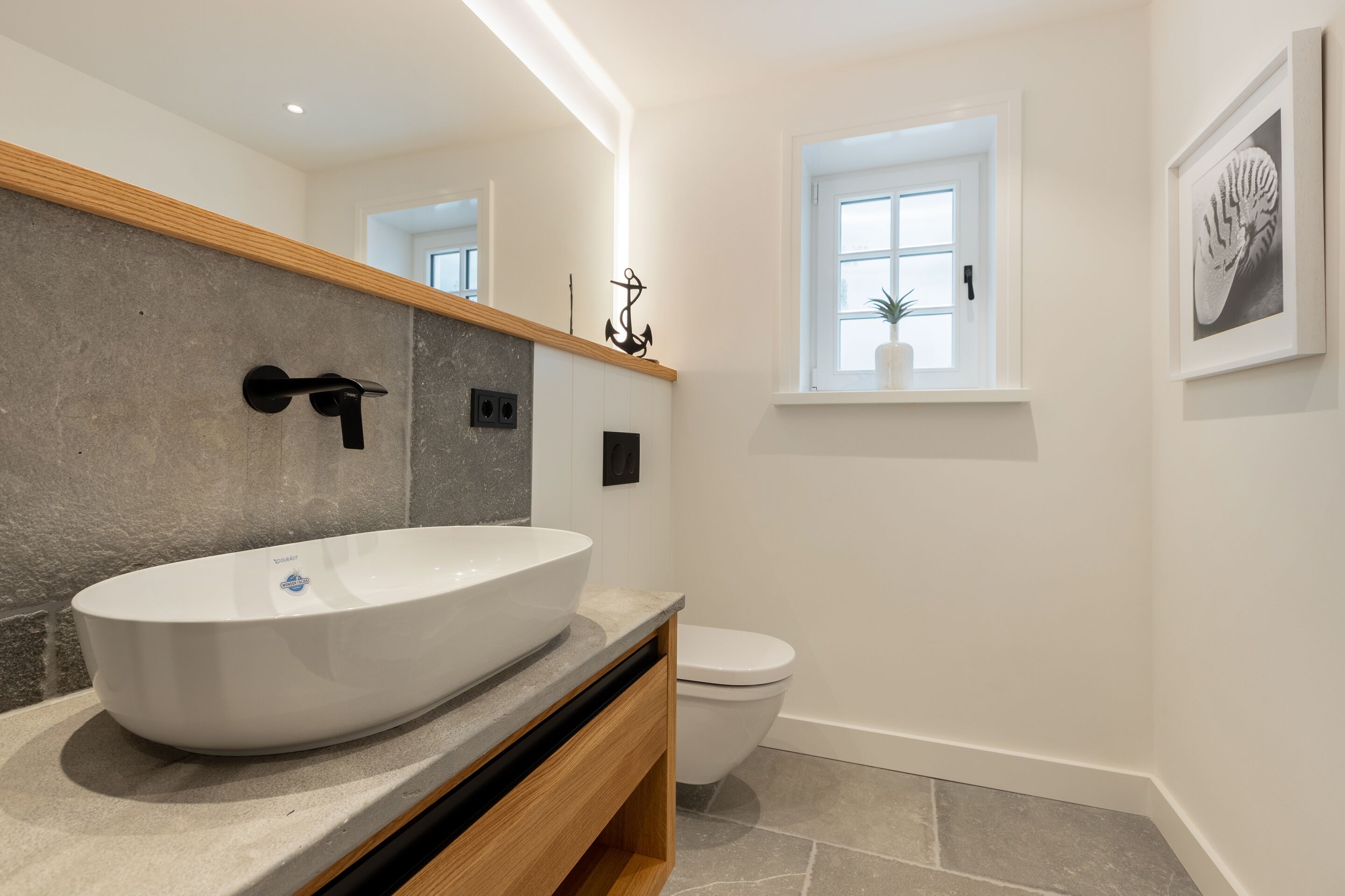 A modern bathroom with a large oval sink, a wooden vanity, a white toilet, and a small window with a plant.