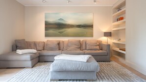 Un salon moderne avec un canapé d’angle, un pouf blanc et un grand tableau représentant un paysage accroché au mur.