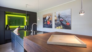 Une cuisine moderne avec un plan de travail en bois, un évier et un crédence éclairée de couleur verte. Deux tableaux ornent le mur et des suspensions lumineuses sont fixées au-dessus du plan de travail.