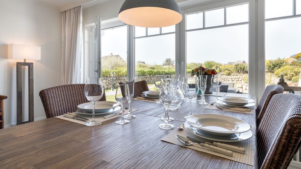 Mesa posta com pratos, copos e talheres, com vista para o exterior através de grandes janelas.