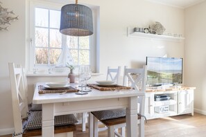 Un coin repas avec une table blanche dressée pour quatre personnes, un téléviseur posé sur un meuble en bois et une fenêtre donnant sur des arbres.
