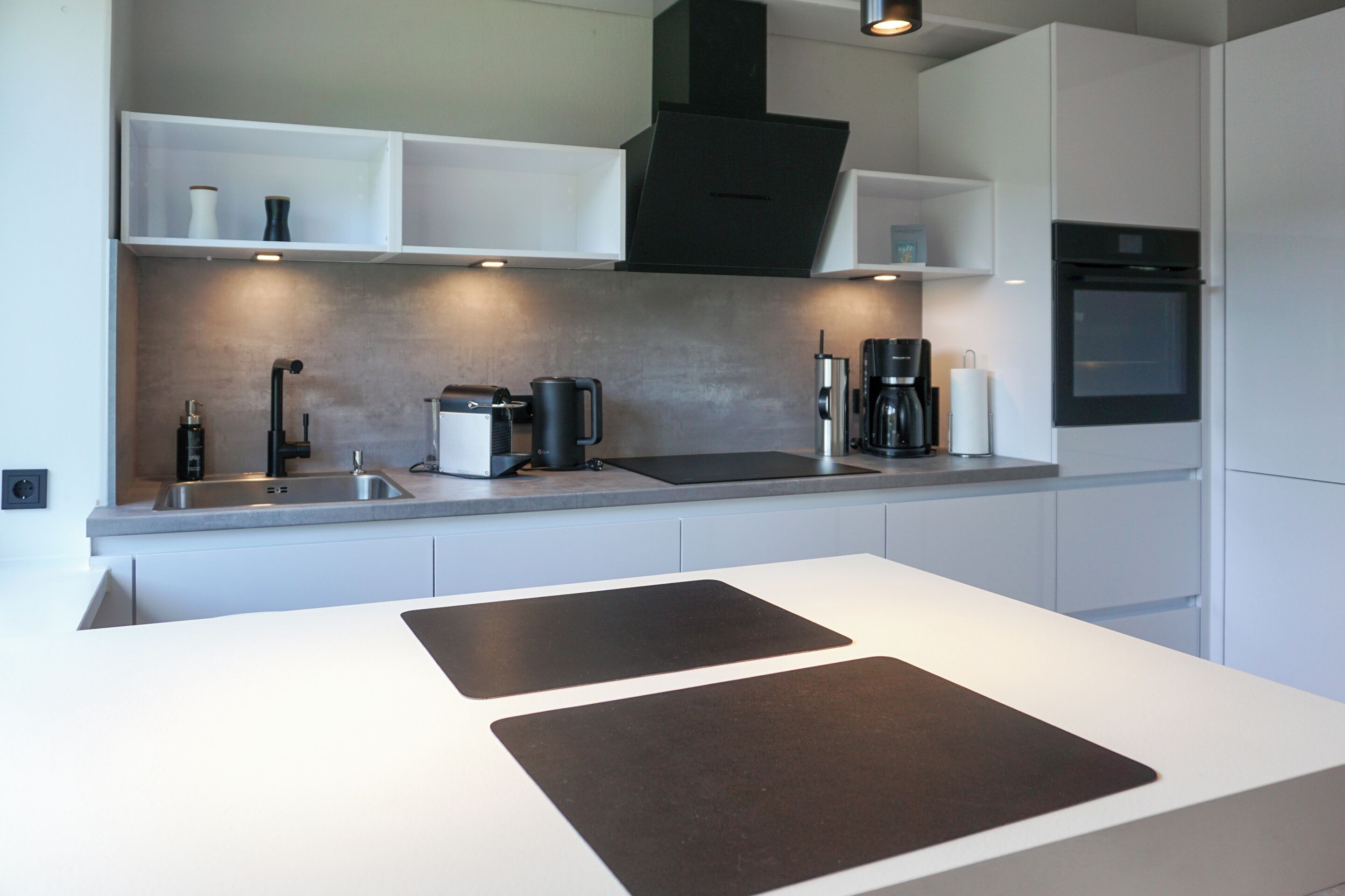 A modern kitchen with white cabinetry, a black range hood, a built-in oven, and a countertop with a coffee maker and kettle.