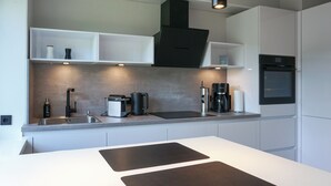 A modern kitchen with white cabinetry, a black range hood, a built-in oven, and a countertop with a coffee maker and kettle. - Warme Farben, ein Hochwertiger Holzboden und Maritim Blaue Accessoires (List auf Sylt)