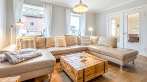 A modern living room with a sectional sofa, a wooden coffee table, and large windows with curtains. - Warme Farben, ein Hochwertiger Holzboden und Maritim Blaue Accessoires (List auf Sylt)