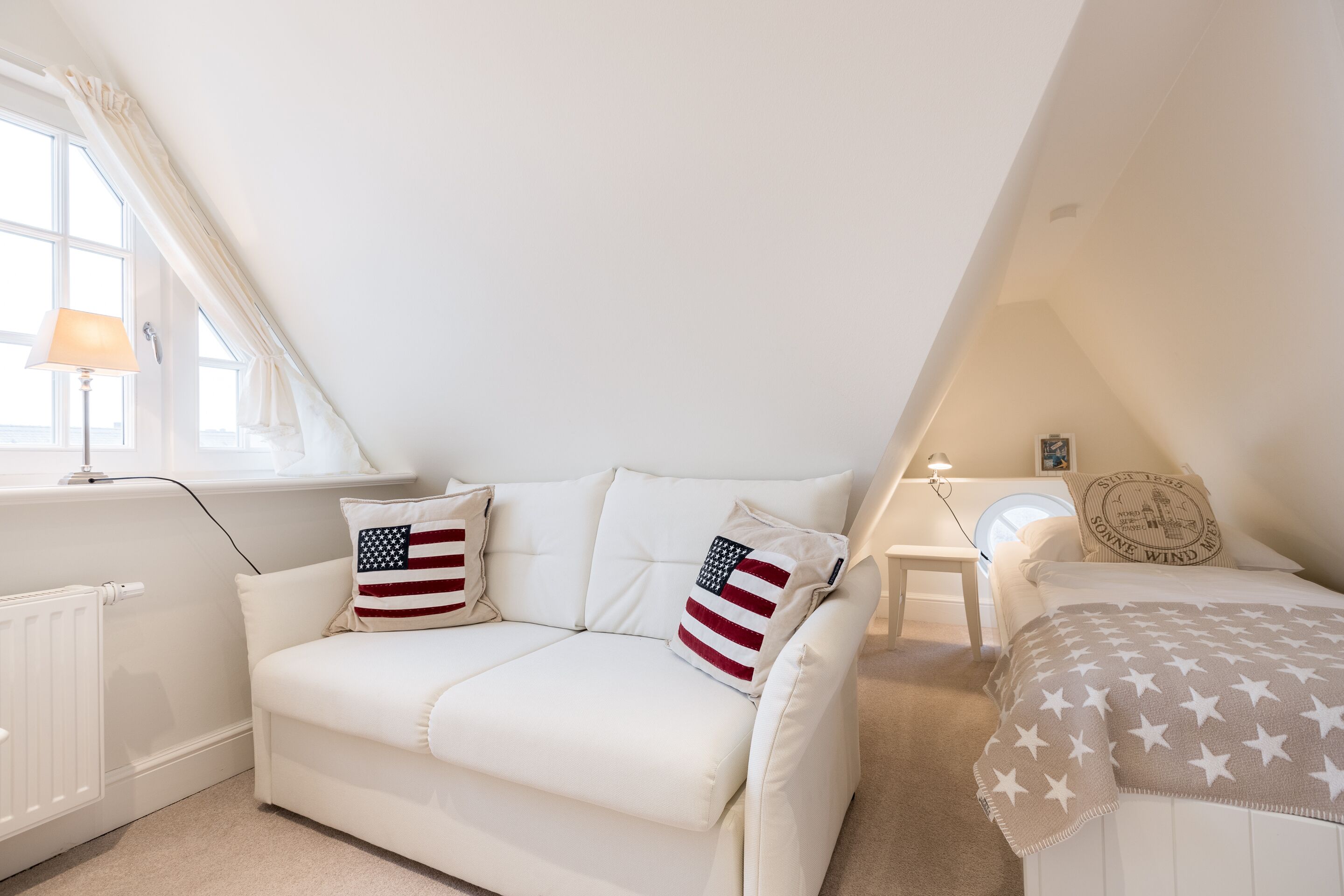 A white, L-shaped sofa with American flag pillows, a white bedside table, a round mirror, and a bed with a star-patterned blanket.