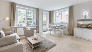 Ein geräumiges Wohnzimmer mit Sofa, Couchtisch und Essbereich. Große Fenster mit Vorhängen, ein Flachbildfernseher und eine Heizung sind ebenfalls vorhanden.