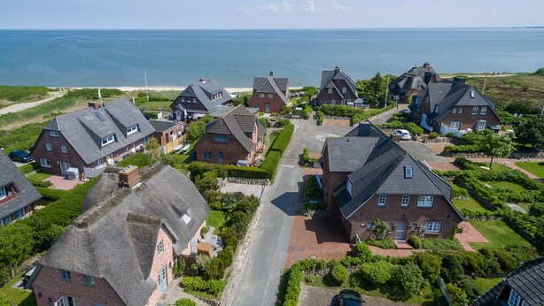 Ein Küstenwohngebiet mit Häusern mit steilen Dächern und Backsteinbauweise, umgeben von viel Grün und Blick aufs Meer.