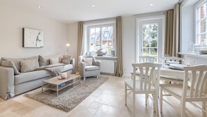 Ein modernes Wohnzimmer mit grauem Sofa, einem kleinen Sessel, einem Esstisch mit Stühlen und großen Fenstern mit Vorhängen.