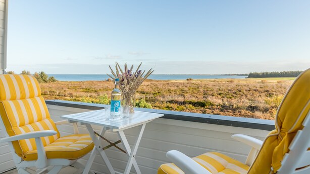 A balcony with two yellow-striped chairs, a small table with a bottle and glasses, and a view of a grassy landscape and water.