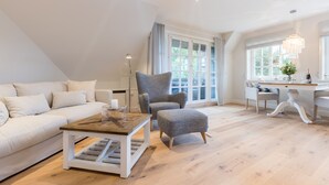 A modern living room with a white sofa, a wooden coffee table, a grey armchair, and a dining area with a white table and chairs.