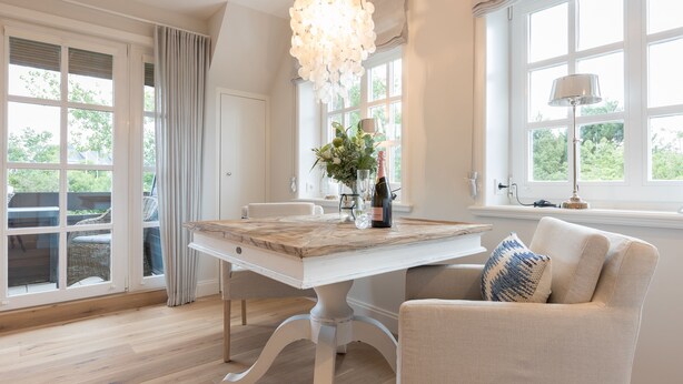 A dining area with a white table, chairs, a chandelier, and a view through large windows.