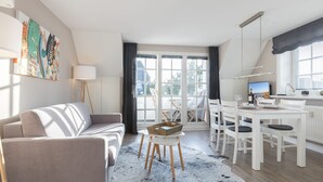 A modern living room with a sofa, dining table, and chairs. There is a large window with curtains, a painting on the wall, and a lamp. - 3-room apartment on the upper floor/ground floor with approx. 54 sqm floor space in Munkmarsch (Sylt)