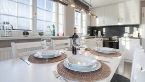 A modern kitchen with white cabinetry, a dining table set for four, and large windows with blinds. - 3-room apartment on the upper floor/ground floor with approx. 54 sqm floor space in Munkmarsch (Sylt)