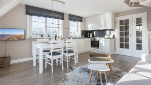 A modern kitchen with white cabinets, a dining area with a white table and chairs, and a large window with a view of the outdoors. - 3-room apartment on the upper floor/ground floor with approx. 54 sqm floor space in Munkmarsch (Sylt)