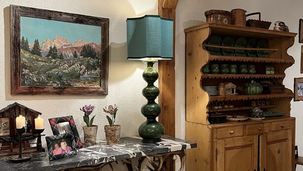 Un interior rústico con una mesa de madera, una lámpara verde, un cuadro enmarcado y un aparador con estantes.