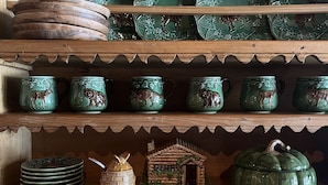 Una estantería de madera con diversos objetos de cerámica, como platos, tazas y un modelo decorativo de una casa.