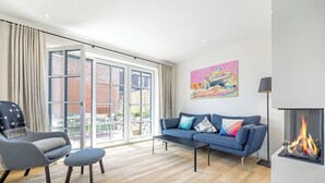 A modern living room with a sofa, armchair, and ottoman, a fireplace, and a large window with a view of a brick building. - Hausteil mit ca. 95qm Nutzfläche im EG, UG, OG und DG (Hörnum (Sylt))