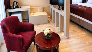 A hotel room with a bed, a TV, a red armchair, a small table with flowers, and a white console table with a kettle. - Ardos Park Hotel (Ankara)